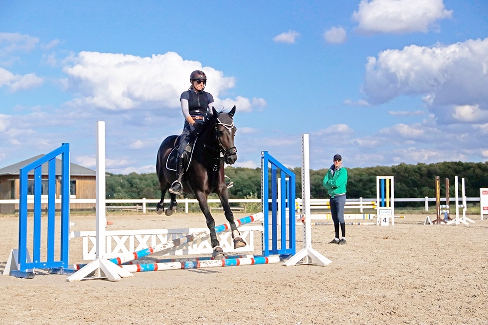 Auf Reitplatz: Reiterin auf dunkelbraunem Pferd überspringt Hindernis, ein Person schaut zu
