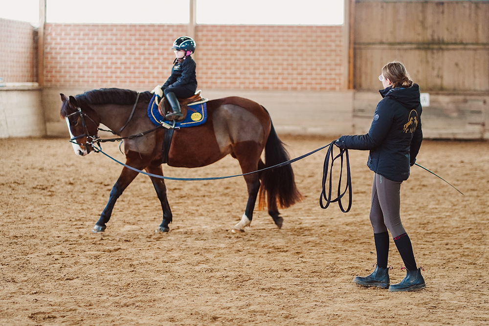 Kind reitet in Reithalle langsam im Kreis, Trainerin hält Pferd an Leine